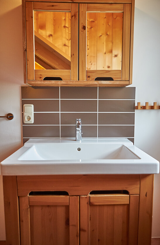 Waschbecken mit Wasserhahn, Holz-Spiegelschrank, braune Fliesen.