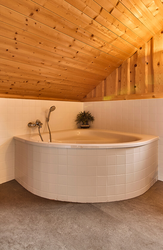 Eckbadewanne, weiße Fliesen, schräge Holzdecke.