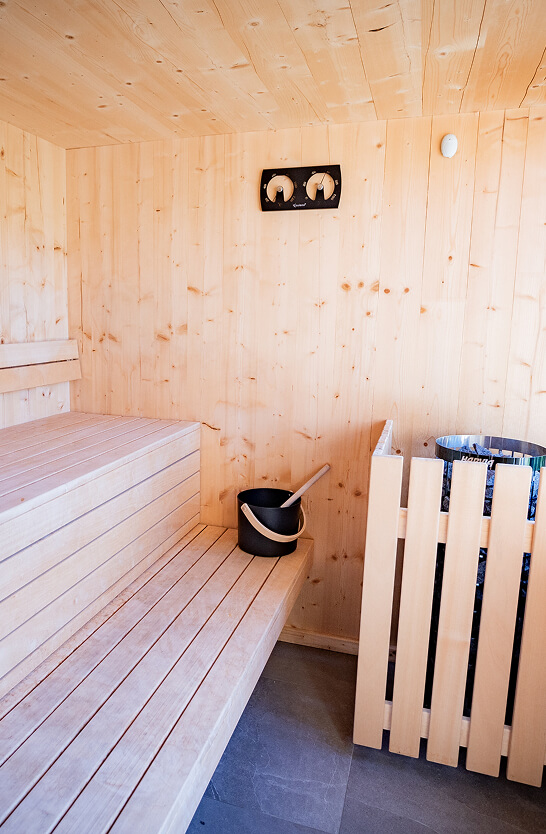 Heller Saunaraum mit Holzbänken, Ofen, Eimer und Thermometer.