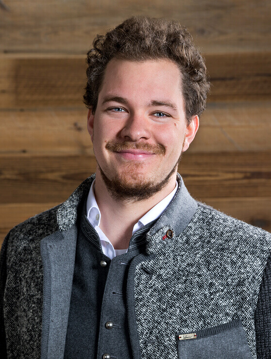 Lächelnder Mann im Trachtenjanker vor Holzwand.