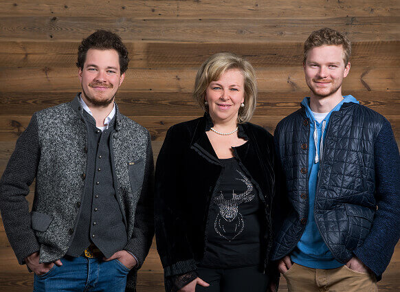 Zwei junge Männer, eine Frau lächeln vor Holzwand.