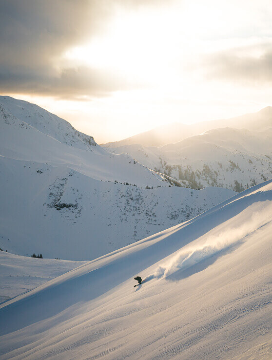 Snowboarder fährt Hang hinab, sprüht Schnee, Abendsonne.