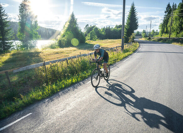 Radfahrer auf Straße am See, lange Schatten bei Sonne.