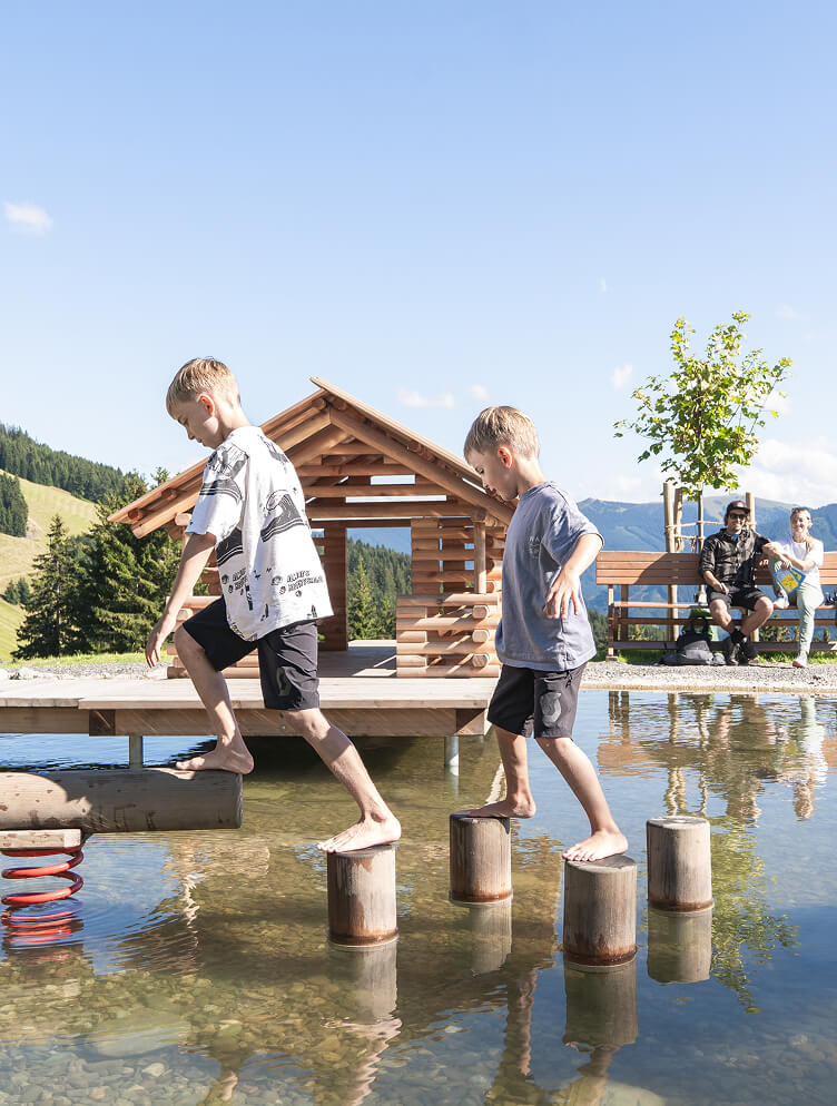 Zwei Jungen balancieren barfuß auf Holzstämmen im Wasser.