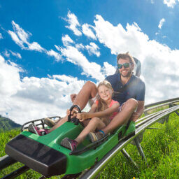 Mann und Mädchen fahren lachend Sommerrodelbahn.