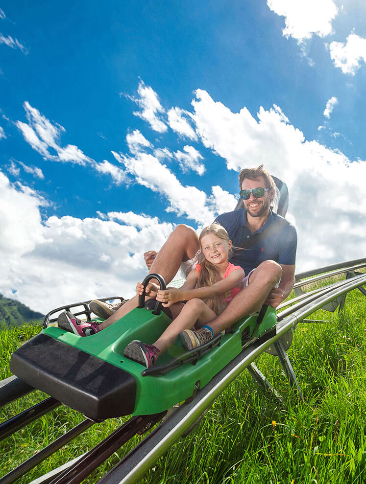 Mann und Mädchen fahren lachend Sommerrodelbahn.