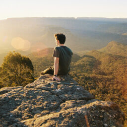 Person sitzt auf Fels, blickt auf bewaldete Berge.