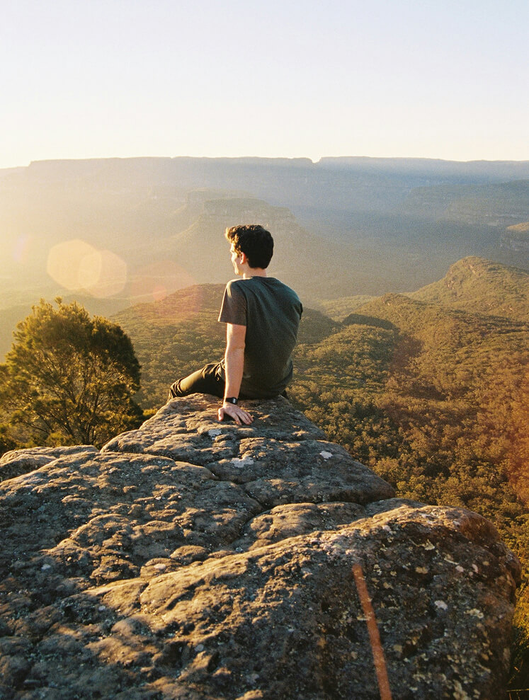 Person sitzt auf Fels, blickt auf bewaldete Berge.