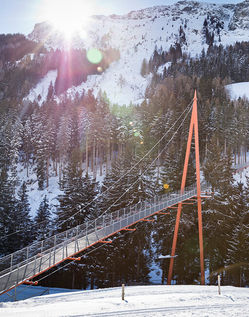 Orange Hängebrücke im verschneiten Bergwald bei Sonne.
