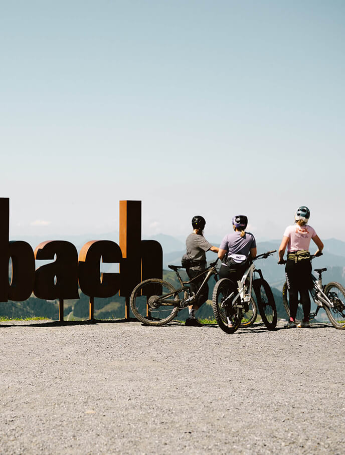 Drei Mountainbikerinnen stehen vor großem Saalbach Schild mit Bergen.